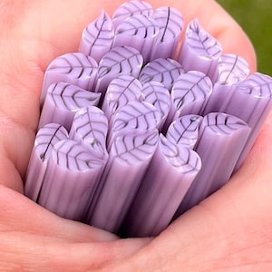 May include: A collection of cylindrical light purple glass canes, each featuring a detailed leaf pattern with darker purple veins running through the center and extending to the edges, arranged in a tight bundle with smooth, rounded ends.
