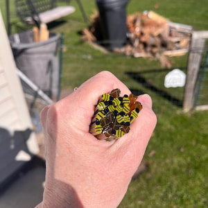 May include: A collection of glass rods, each featuring a yellow and black bee pattern. The rods are held in a hand, with a silver bracelet on the wrist. The background shows a sunny outdoor scene.