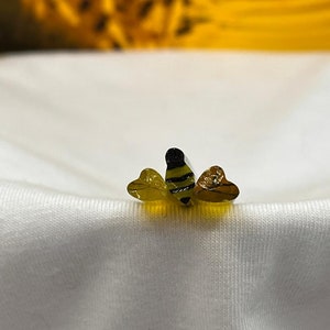 May include: A small, yellow and black bee charm with a three-dimensional design. The bee is sitting on a white fabric surface.