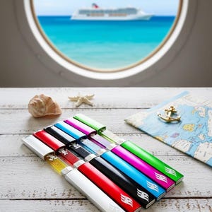May include: A collection of colorful, rectangular lighters with silver accents, arranged on a white wooden surface. The lighters are in various colors, including red, black, blue, and green. A map, seashell, and starfish are also visible, with a cruise ship in the background.