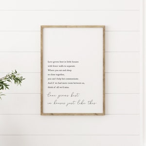 May include: A framed print with a light wood frame displays a poem in black script. The poem begins, "Love grows best in little houses..." and ends with "love grows best in houses just like this." The print hangs on a white shiplap wall.