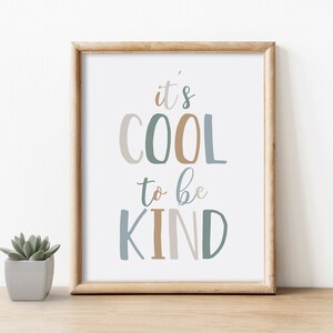 May include: A framed print with the words "it's COOL to be KIND" in various colors. The frame is light brown, and a small succulent in a gray pot sits to the left of the frame. The text is in a playful font.