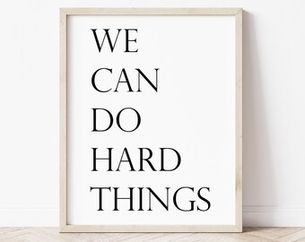 May include: A framed print with the words "WE CAN DO HARD THINGS" in a bold, black font. The text is centered on a white background, encased in a light-colored wooden frame. The minimalist design is suitable for various decor styles.