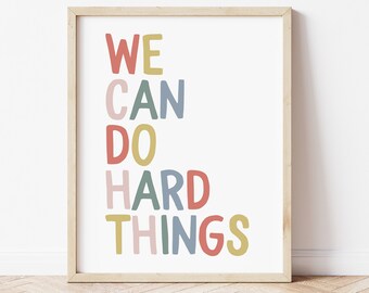 May include: A framed print with the words "WE CAN DO HARD THINGS" in colorful, playful lettering. The text is arranged vertically on a white background, framed by a light wood border. The artwork is displayed against a white wall.