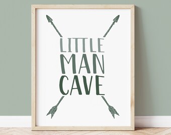 May include: A framed print with the words "LITTLE MAN CAVE" in green and gray lettering, with two arrows pointing towards the text. The print has a light wood frame and is displayed against a light green wall.
