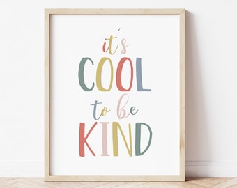 May include: A framed art print with the words "it's COOL to be KIND" in colorful, playful lettering. The print has a light wood frame and is displayed against a white wall, with a wooden floor visible below.