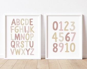 May include: Two framed prints with white borders. One displays the alphabet in pink, beige, and light brown. The other shows numbers 0-10 in the same colors. The prints are displayed on a light wood floor.