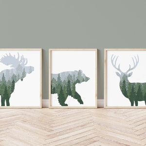 May include: Three framed art prints featuring silhouettes of a moose, bear, and deer, each filled with a forest scene. The animals are in shades of green and gray, set against a white background, with light wood frames.