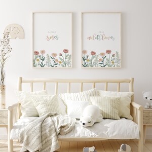 May include: Two framed floral art prints with the phrases "In a field of roses" and "She is a wildflower." The prints feature colorful flowers at the bottom. The prints hang above a light wood daybed with pillows and a blanket.