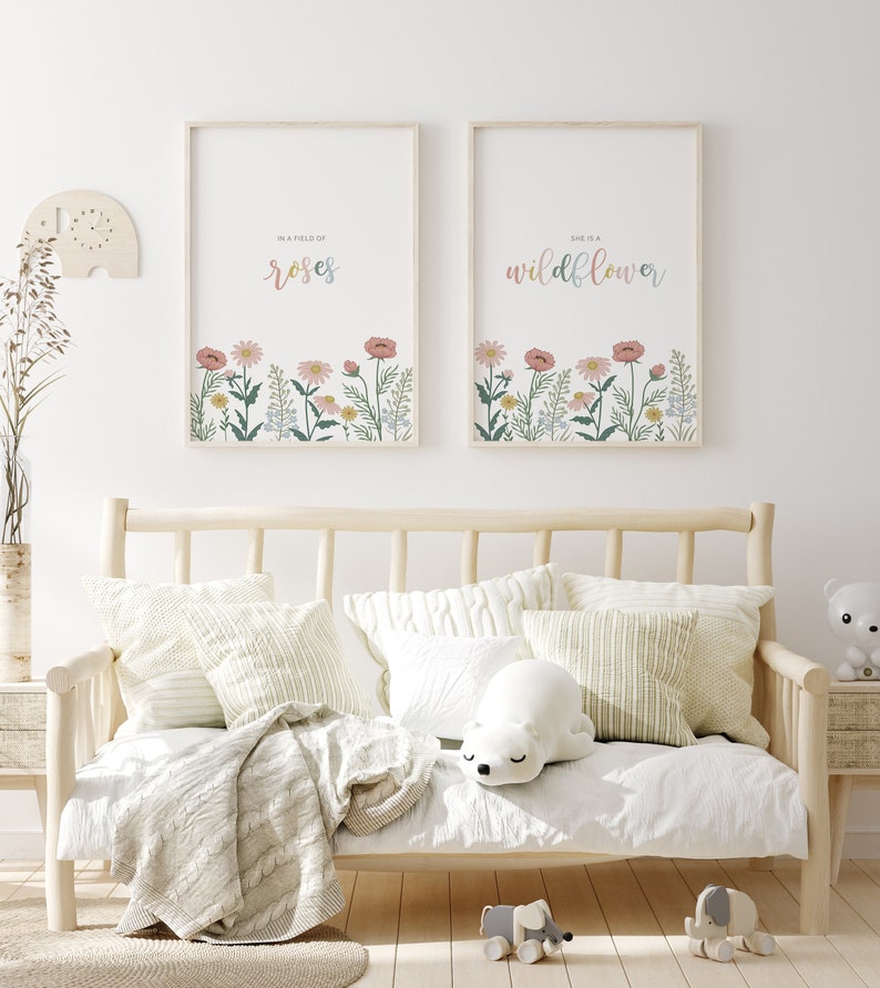 May include: Two framed floral art prints with the phrases "In a field of roses" and "She is a wildflower." The prints feature colorful flowers at the bottom. The prints hang above a light wood daybed with pillows and a blanket.