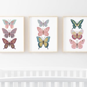May include: Three framed butterfly art prints. Each print features three colorful butterfly illustrations in shades of pink, blue, yellow, and green. The artwork is displayed on a white background with a light wood frame.