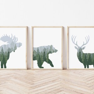 May include: Three framed art prints featuring silhouettes of a moose, bear, and deer. Each animal is filled with a forest scene in shades of green and gray. The frames are light brown, and the background is white.