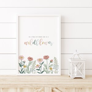 May include: A framed print with the text "In a field of roses she is a wildflower." The print features a colorful floral border with pink, yellow, and blue flowers. A white lantern sits to the right of the framed print.