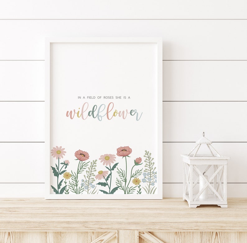 May include: A framed print with the text "In a field of roses she is a wildflower." The print features a colorful floral border with pink, yellow, and blue flowers. A white lantern sits to the right of the framed print.