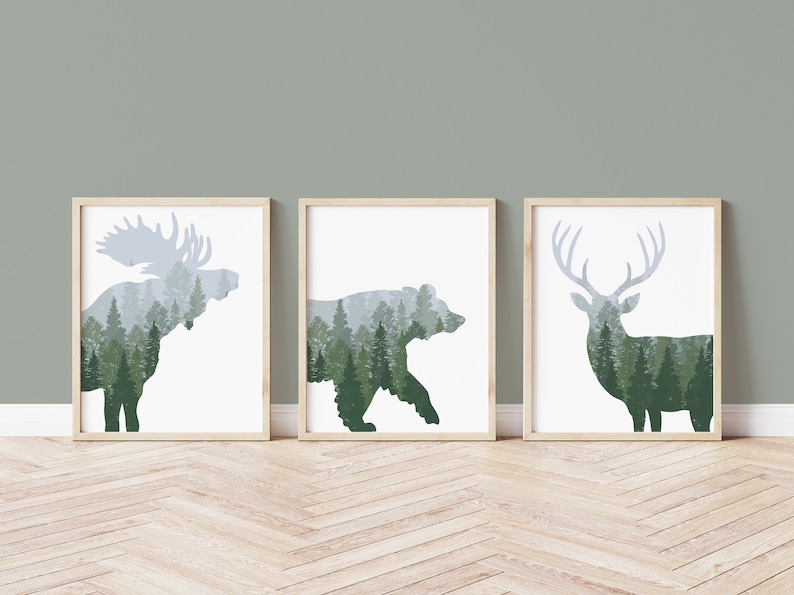 May include: Three framed art prints featuring silhouettes of a moose, bear, and deer, each filled with a forest scene. The animals are in shades of green and gray, set against a white background, with light wood frames.