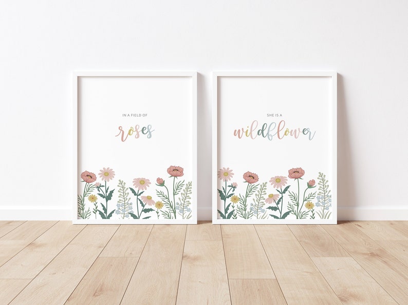 May include: Two framed art prints with white borders. The left print reads "IN A FIELD OF roses" in pastel colors, above a floral border. The right print reads "SHE IS A wildflower" in pastel colors, above a floral border.
