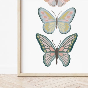 May include: A framed print featuring three colorful butterfly illustrations. The butterflies have detailed wing patterns in shades of pink, blue, yellow, and green. The artwork is set against a white background, and the frame is a light wood color.