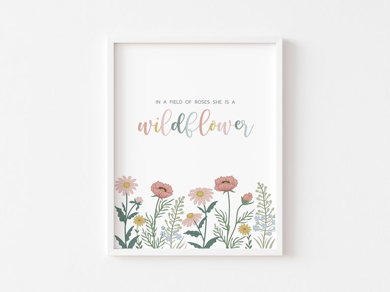 May include: White framed art print featuring the text "IN A FIELD OF ROSES SHE IS A wildflower" in pastel colors. The bottom of the print displays a colorful illustration of wildflowers in pink, yellow, and blue.