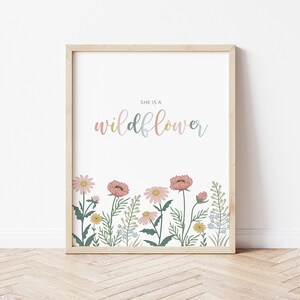 May include: A framed art print featuring the text "SHE IS A wildflower" in pastel colors. The bottom of the print displays a colorful illustration of wildflowers, including pink, yellow, and blue flowers, with green foliage.