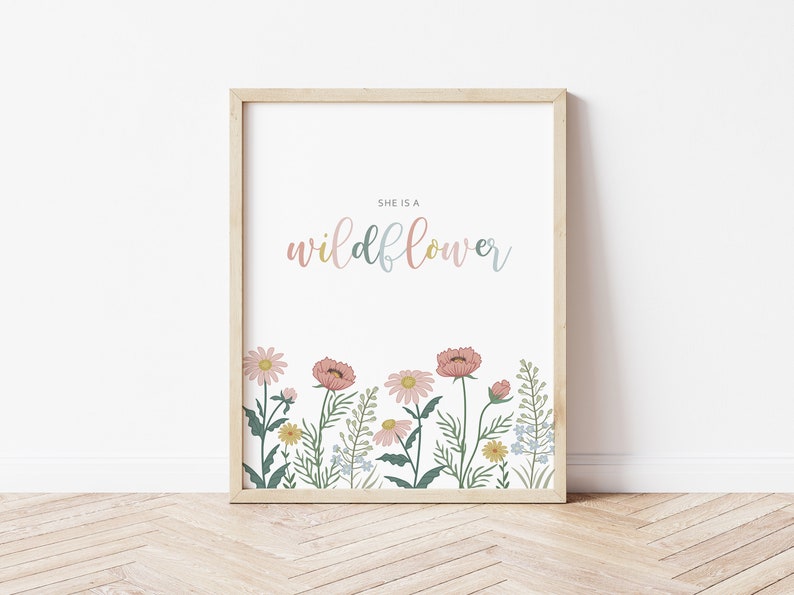 May include: A framed art print featuring the text "SHE IS A wildflower" in pastel colors. The bottom of the print displays a colorful illustration of wildflowers, including pink, yellow, and blue flowers, with green foliage.