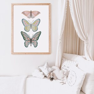May include: A framed print featuring three colorful butterfly illustrations. The butterflies are in shades of pink, blue, yellow, and green, set against a white background. The frame is made of light-colored wood. The artwork is displayed in a child's bedroom.