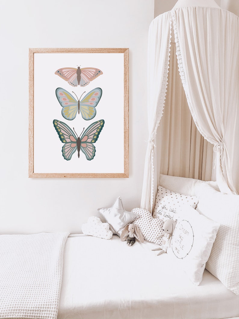 May include: A framed print featuring three colorful butterfly illustrations. The butterflies are in shades of pink, blue, yellow, and green, set against a white background. The frame is made of light-colored wood. The artwork is displayed in a child's bedroom.