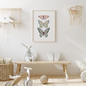 May include: A framed print featuring three colorful butterfly illustrations. The butterflies are in shades of pink, blue, yellow, and green, set against a white background. The artwork is displayed in a light-colored wooden frame, suitable for a nursery or children's room.