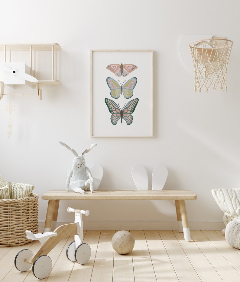 May include: A framed print featuring three colorful butterfly illustrations. The butterflies are in shades of pink, blue, yellow, and green, set against a white background. The artwork is displayed in a light-colored wooden frame, suitable for a nursery or children's room.