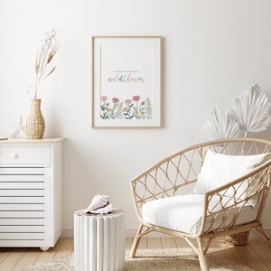 May include: A framed print with the text "In a field of roses she is a wildflower" above a row of colorful flowers. The print is displayed in a light-colored wooden frame. The room also features a woven chair, a white cabinet, and a decorative vase.