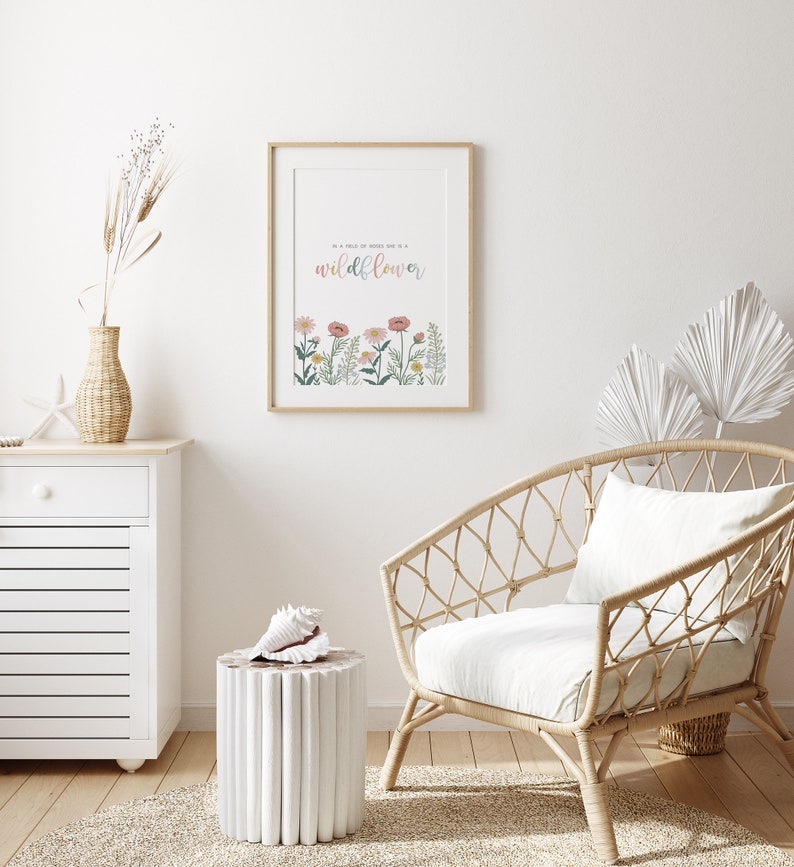 May include: A framed print with the text "In a field of roses she is a wildflower" above a row of colorful flowers. The print is displayed in a light-colored wooden frame. The room also features a woven chair, a white cabinet, and a decorative vase.