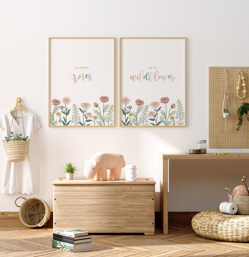 May include: Two framed art prints with floral designs and text. One reads "In a field of roses" and the other "She is a wildflower." The prints feature a pastel color palette and are displayed above a wooden chest.