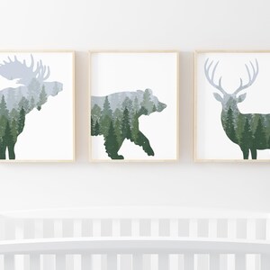 May include: Set of three framed art prints featuring silhouettes of a moose, bear, and deer. Each animal is filled with a forest scene in shades of green and gray. The frames are light wood, and the background is white.