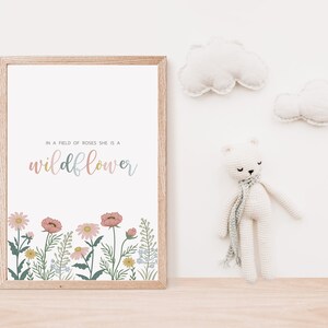 May include: A framed print with the text "In a field of roses she is a wildflower" in pastel colors. The bottom of the print features a floral border with pink, yellow, and blue flowers. A white crocheted bear with a scarf and two cloud-shaped wall decorations are also visible.