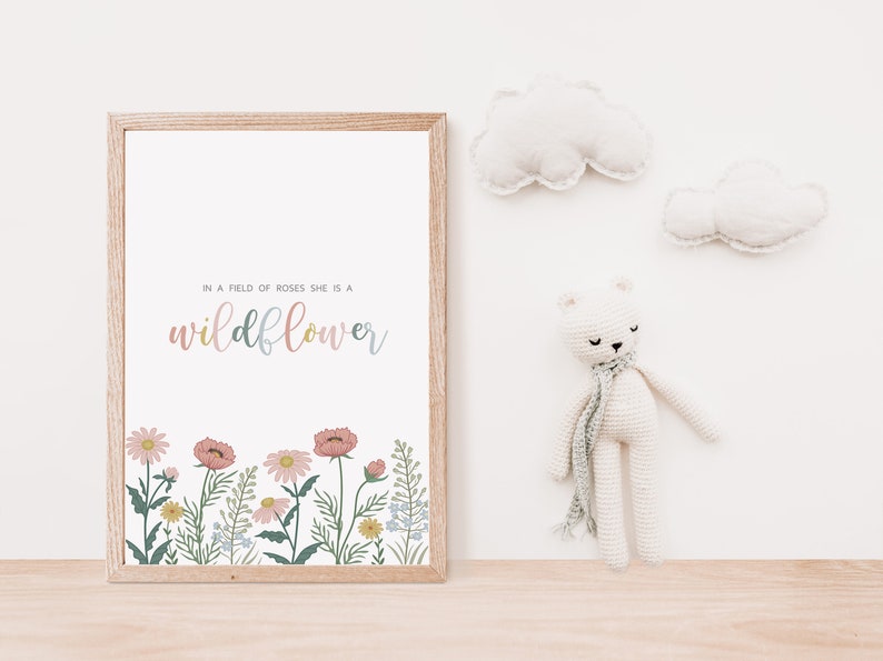 May include: A framed print with the text "In a field of roses she is a wildflower" in pastel colors. The bottom of the print features a floral border with pink, yellow, and blue flowers. A white crocheted bear with a scarf and two cloud-shaped wall decorations are also visible.