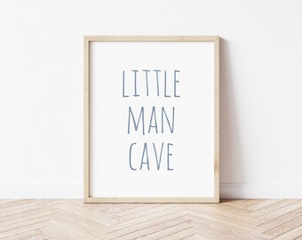 May include: A framed print with the words "LITTLE MAN CAVE" in blue lettering. The print is in a light wood frame and is displayed against a white wall. The floor has a light wood pattern.