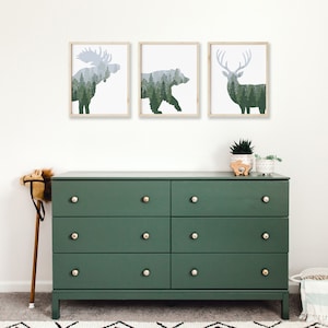 May include: Three framed art prints featuring silhouettes of a moose, bear, and deer filled with a forest scene. Below, a dark green six-drawer dresser with gold knobs. A toy horse and decorative plants complete the room.