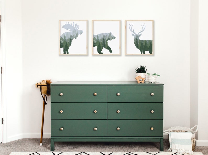May include: Three framed art prints featuring silhouettes of a moose, bear, and deer filled with a forest scene. Below, a dark green six-drawer dresser with gold knobs. A toy horse and decorative plants complete the room.