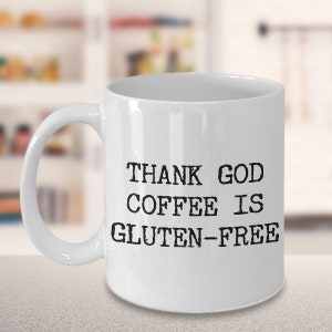 Puede incluir: Taza de caf&eacute; de cer&aacute;mica blanca con texto negro que dice "THANK GOD COFFEE IS GLUTEN-FREE".