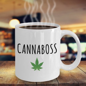 May include: White ceramic coffee mug with a black graphic that says "CANNABOSS" and a green marijuana leaf design.