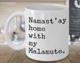 May include: White ceramic mug with the text "Namast'ay home with my Malamute" in black. The mug has a curved handle and is set against a blurred background of a kitchen.