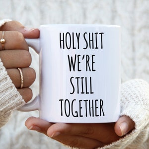 May include: White ceramic coffee mug with black text that reads "HOLY SHIT WE'RE STILL TOGETHER".