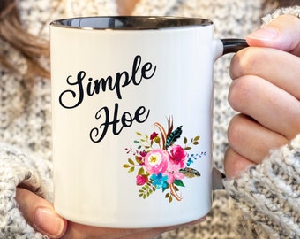 May include: White ceramic mug with a black rim and handle. The mug has a floral design and the text "Simple Hoe" in black cursive lettering.