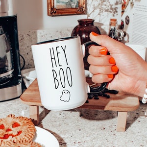 May include: White ceramic coffee mug with black text that reads "Hey Boo" and a small ghost illustration. The mug has a black rim and handle.