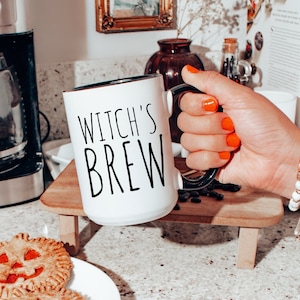 May include: White ceramic mug with a black rim and the text "WITCH'S BREW" in black lettering. The mug is being held by a hand with orange nail polish.
