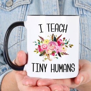 Peut inclure: Une tasse &agrave; caf&eacute; blanche avec une anse noire. La tasse est d&eacute;cor&eacute;e d'un motif floral et du texte "J'enseigne les petits humains".