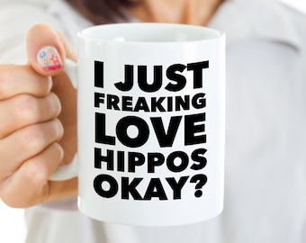 May include: White ceramic mug with the text "I JUST FREAKING LOVE HIPPOS OKAY?" in bold black letters. The mug is held by a person with a light complexion. The mug is a standard size for coffee or tea.