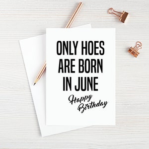 May include: A white card with black text that reads "ONLY HOES ARE BORN IN JUNE" and "Happy Birthday" in cursive. The card is on a white wooden surface with a pencil and two gold paper clips.