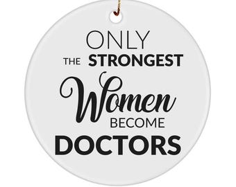 Puede incluir: Un adorno redondo de cerámica blanca con texto negro que dice ONLY THE STRONGEST Women BECOME DOCTORS. El adorno tiene un pequeño orificio en la parte superior para colgarlo y está suspendido por un hilo dorado fino sobre un fondo blanco.