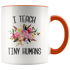 Peut inclure: Une tasse &agrave; caf&eacute; en c&eacute;ramique blanche avec un bord et une poign&eacute;e orange. La tasse pr&eacute;sente un motif floral et le texte "J'enseigne de petits humains".