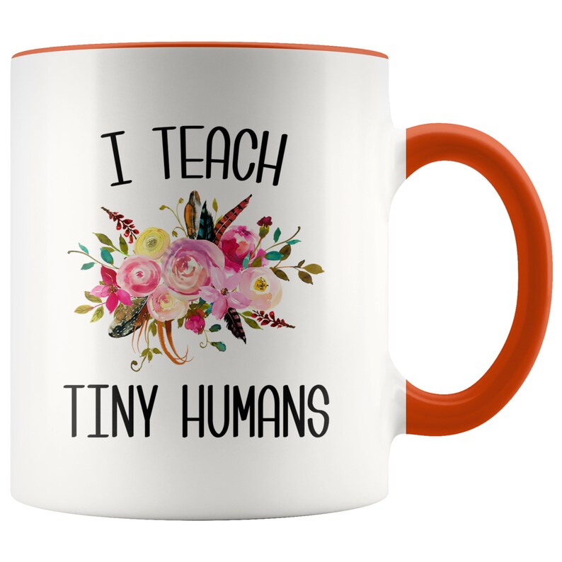 Peut inclure: Une tasse &agrave; caf&eacute; en c&eacute;ramique blanche avec un bord et une poign&eacute;e orange. La tasse pr&eacute;sente un motif floral et le texte "J'enseigne de petits humains".